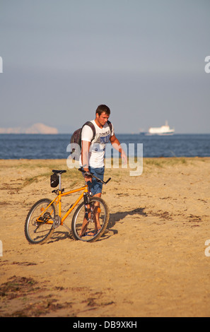 Hübscher junger Mann schob Fahrrad entlang Studland Beach mit Schwerpunkt Boot und Klippen im Hintergrund auf sonnigen Juliabend Stockfoto