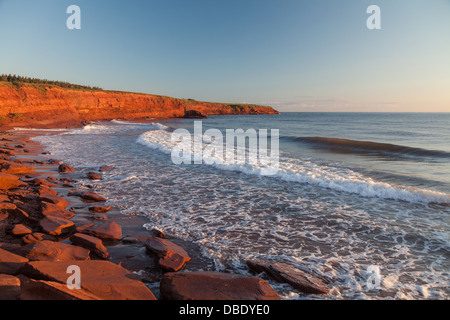 Die felsige Küste von Prince Edward Island bei Tagesanbruch beleuchtet die Klippen und Felsen leuchtend rot. Stockfoto