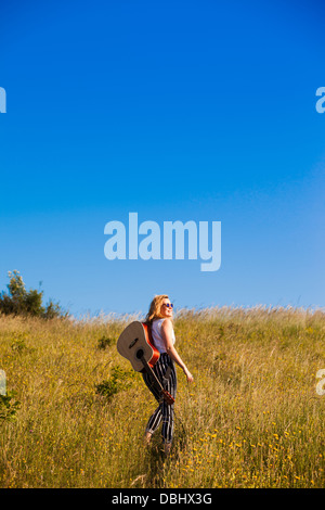 Teenager-Mädchen zu Fuß durch eine Wiese mit einer Gitarre. Im Freien. Sommer. Stockfoto