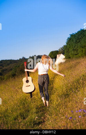 Teenager-Mädchen zu Fuß durch eine Wiese mit einer Gitarre. Im Freien. Sommer. Stockfoto