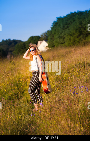 Teenager-Mädchen zu Fuß durch eine Wiese mit einer Gitarre. Im Freien. Sommer. Stockfoto