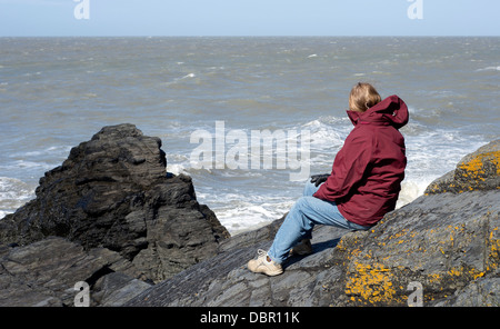 Eine Frau-Website auf einem Felsen mit Blick auf das Meer in der Nähe von Tresaith Strand im Südwesten von Wales, an einem sonnigen, aber lebhaften Tag. Stockfoto