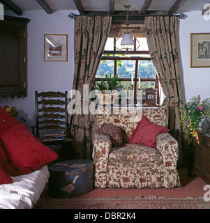 Gemusterte Vorhänge am Fenster über florale Sessel im Ferienhaus Wohnzimmer mit antiken Ladder-back Stuhl und bemalte Runde box Stockfoto