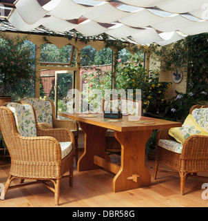 Korbsessel und einfachen Holztisch im Wintergarten mit weißen Decke Jalousien und Blick auf den Garten Stockfoto