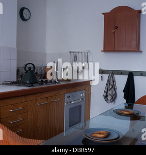 Kleine hölzerne Wand Schrank und Glas Esstisch in moderne Küche mit Edelstahl-Backofen-Set aus hellem Holz ausgestattet Einheit Stockfoto