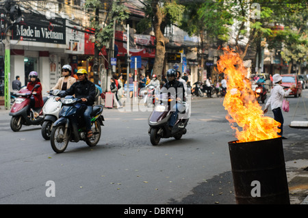 HANOI, Vietnam – Eine 44-Liter-Trommel brennt mit hohen Flammen am Straßenrand, während Motorroller in der Altstadt von Hanoi vorbeieilen. Stockfoto