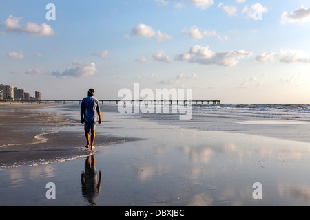 Mann zu Fuß am Strand am Rande der Brandung in der Morgendämmerung mit Angelsteg darüber hinaus sichtbar. Stockfoto