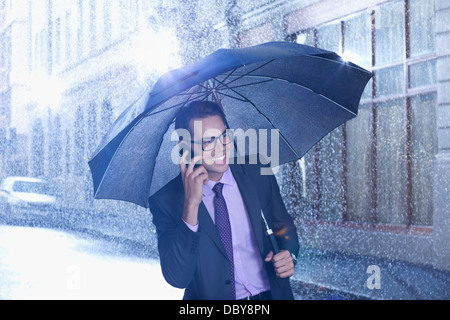 Glücklich Kaufmann reden über Handy unter Dach im verregneten Straße Stockfoto