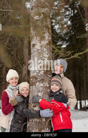 Porträt der glückliche Familie umarmt Baumstamm im verschneiten Wald Stockfoto