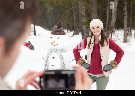 Mann fotografiert lächelnde Frau mit Händen auf den Hüften neben Schneemann im Wald Stockfoto