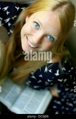 eine junge, attraktive Frau sitzen auf einer Couch, einem Buch schaut und lächelt. Stockfoto