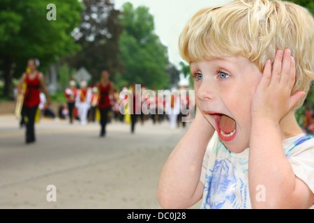 Ein niedlichen Kleinkind Jungen deckt seine Ohren, wie er eine Schule marching Band Spaziergang in einer Parade Uhren. Stockfoto