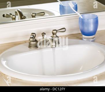 Foto von weißen Badezimmer Waschbecken und laufenden Wasserhahn mit blauen Tasse, Zahnbürste und Spiegel im Hintergrund Stockfoto