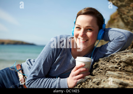 Porträt der jungen Frau an Küste mit Kaffee und Kopfhörer Stockfoto