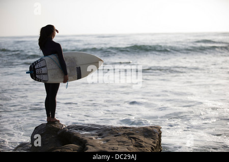 Junge Frau, die auf Felsen mit Surfbrett stehend Stockfoto