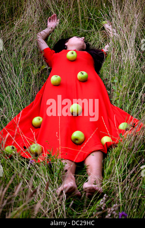 eine Frau in einem roten Kleid, liegt auf der Wiese mit grünen Äpfeln an ihrem Kleid Stockfoto