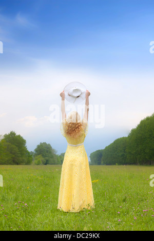 eine blonde Frau in einem gelben Kleid auf einer Wiese, hält einen Sonnenhut Stockfoto
