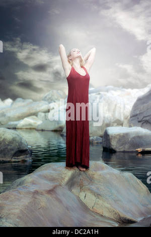 eine Frau in einem roten Kleid steht auf einem Felsen im Wasser und die Sonne genießen Stockfoto