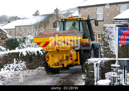 Ein Traktor mit einer Körnung Verbreitung Maschine ausgestattet Antriebe durch eine kleine verschneite Straße in Askrigg, North Yorkshire Stockfoto