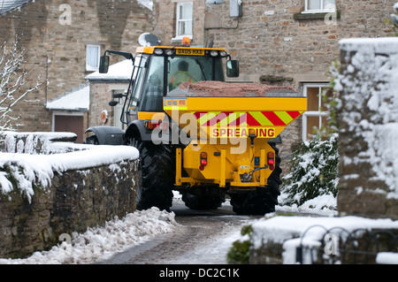 Ein Traktor mit einer Körnung Verbreitung Maschine ausgestattet Antriebe durch eine kleine verschneite Straße in Askrigg, North Yorkshire Stockfoto