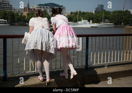 Zwei Mädchen Kleider tragen Lolita schlanke über die Schranke über die Themse auf der South Bank, London, UK. Lolita Mode ist eine Subkultur mit Ursprung in Japan, die auf viktorianische Kleidung basiert. Das Aussehen begann auch in erster Linie als eine der Bescheidenheit mit einem Fokus auf Qualität mit einem Gefühl der Gotik. Die ursprüngliche Form ist einem knielangen Rock mit einer "Cupcake" Form von Unterröcken unterstützt, aber hat zu verschiedenen Arten von Kleidungsstücken erweitert. Blusen, kniehohen Socken oder Strümpfe und Kopfschmuck auch getragen. Stockfoto