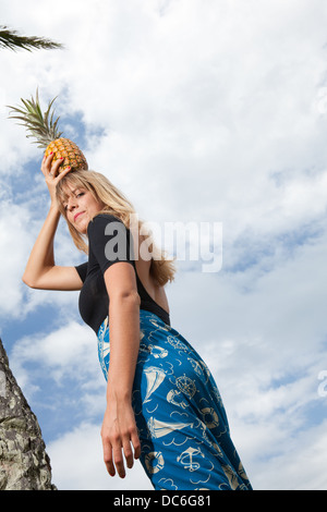 Eine blonde Frau Caucasion steht unter einer Palme mit Ananas auf dem Kopf. Stockfoto