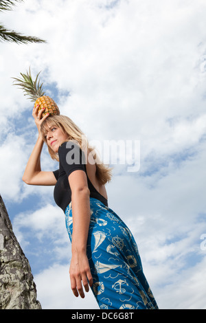 Eine blonde Frau Caucasion steht unter einer Palme mit Ananas auf dem Kopf. Stockfoto