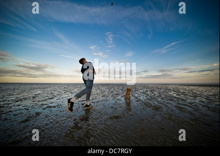 Junger Mann wirft einen Ball für seinen Hund auf Worthing Strand bei Ebbe bei Sonnenuntergang. Stockfoto