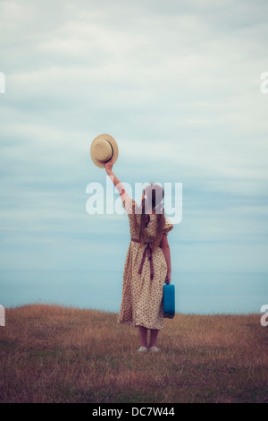 ein Mädchen in ein Vintage-Kleid mit Koffer hält einen Hut Stockfoto