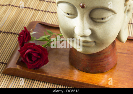 Eine Buddha-Kopf mit zwei Rosen, gelegt auf einer Matte von Stöcken Stockfoto