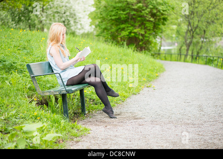 Eine junge attraktive Frau sitzen auf Bank in einem Park in ein Tagebuch zu schreiben Stockfoto