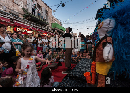 Jerusalem.A Mädchen spielt mit Puppen, als Jongleur im Hintergrund während eines Festes auf dem berühmten "Mahane Yehuda" Markt führt Stockfoto