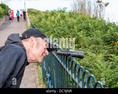 Senior Mann schaut durch dekorative Metall Fernglas auf dekorativen Eisenzaun vor der Klippe - Bournemouth, Dorset Stockfoto