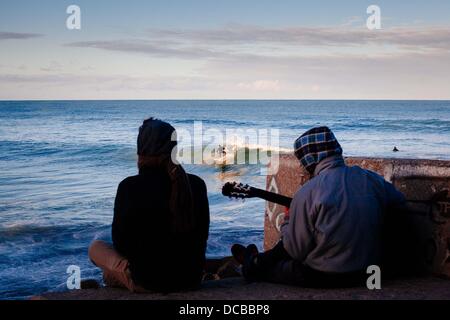 Mar del Plata, Buenos Aires, Argentinien. 14. August 2013. Zwei Jugendliche sitzen und spielen Gitarre wie eine Surfer eine Welle bei Sonnenuntergang fängt. Mar Del Plata ist Argentiniens führende Surfspot sowie einer der beliebtesten Resort Städte Südamerikas. (Kredit-Bild: © Ryan Noble/ZUMA Draht) Stockfoto