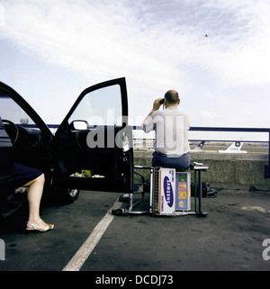 Ein Luftfahrt-Enthusiasten Mann sitzt auf einem umgestürzten Gepäck Wagen auf dem Dach des Terminals am Flughafen London Heathrow, während seine gelangweilte Gattin in ihrem Auto sitzt wartet auf die Heimreise. Stockfoto