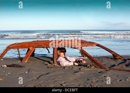 Mann sitzt auf verrosteten Autowrack vergraben im Sand am Strand Stockfoto