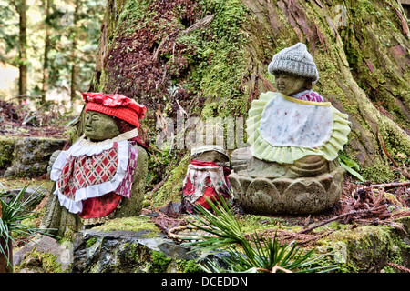 Drei Stein-Jizo-Statuen an der moosbedeckten Basis eines Zedernbaums hinterlassen. Alle sind gehuttet und mit einem Lätzchen ausgestattet. Der alte japanische Okunoin Friedhof. Stockfoto