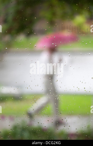 Mann, der einen rosa Regenschirm trägt, der während des Frühjahrsregensturms auf dem Bürgersteig durch Regentropfen auf dem Fensterglas des Hauses läuft Stockfoto