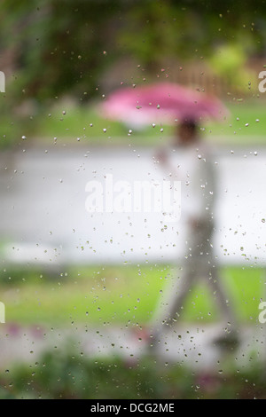 Mann, der einen rosa Regenschirm trägt, der während des Frühjahrsregensturms durch Regentropfen auf dem Glasfenster auf dem Bürgersteig läuft Stockfoto