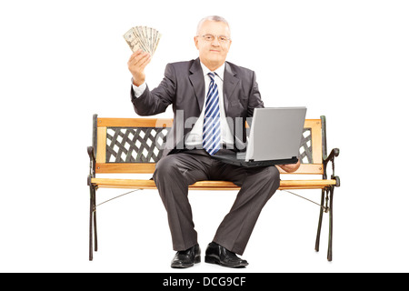 Glücklich reifer Geschäftsmann mit Laptop sitzt auf einer Holzbank und hält US-Dollar Stockfoto