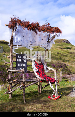 Ich wartete auf den perfekten Mann, einsames Skelett in einem roten Kleid, saß auf einer aufgerüsteten Holzbank aus recyceltem Holz. Carbost, Isle of Skye, Schottland, Großbritannien Stockfoto