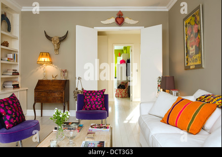 Weißen Sofa von Andrew Morton mit orange Kissen im Wohnzimmer mit lila Samtstühlen und ungewöhnliche Wanddekorationen Stockfoto