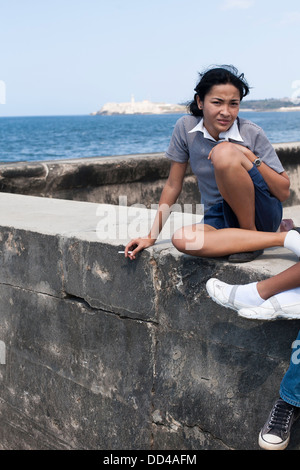 Eine Mädchen in Schuluniform sitzt und raucht eine Zigarette auf dem Deich, aus denen sich die Malecon an der Küste von Havanna, Kuba Stockfoto