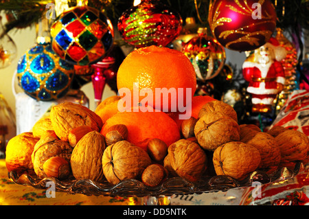 Glas Platte Stand garniert mit Walnüssen, Haselnüssen und Mandarinen mit Weihnachten präsentiert unter Baum nach hinten. Stockfoto