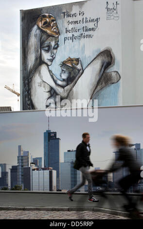 Die Leute gehen vorbei an einem Bauzaun zeigt die Skyline von Frankfurt, während ein neues Wandbild an der Fassade eines Telekom-Gebäudes in Frankfurt Main, Deutschland, 27. August 2013 im Hintergrund gesehen werden kann. Das 15 Meter hohe Kunstwerk des deutschen Künstlerduos "Herakut" ist Teil ihrer "Riesenprojekt Storybook". Im Rahmen des Projekts haben die Künstler besprüht einseitig ein Kinderbuch auf verschiedenen Wänden weltweit und fotografierte ihn. Foto: FRANK RUMPENHORST Stockfoto