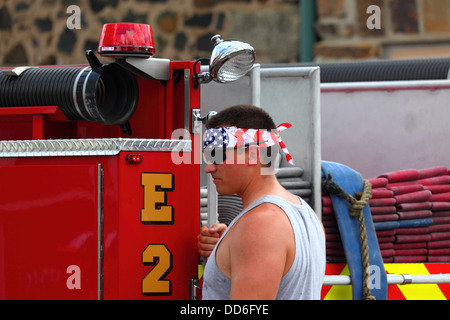 Junger Feuerwehrmann mit einem Stirnband in Stars and Stripes, der auf der Rückseite des Feuerwehrwagens während der Paraden zum 4. Juli in Catonsville, Maryland, USA, fährt Stockfoto