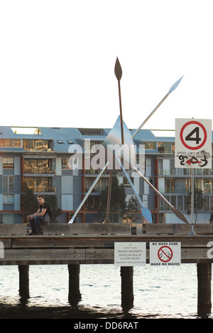 Ein junger Mann sitzt auf einem Hafen Kai in Pyrmont Sydney, als neben einem Edelstahl Skulptur der SubWharyen bezeichnet. Stockfoto