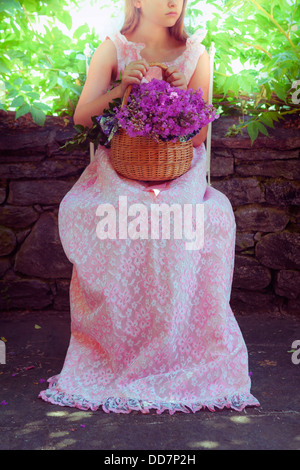 ein Mädchen im rosa Kleid draußen sitzen auf einem Stuhl mit einem Korb voller Wildblumen Stockfoto