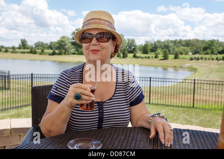Stilvolle senior Frau in einen Hut und eine Sonnenbrille, einen Drink im Freien sitzen an einem Tisch mit Blick auf einen See Stockfoto