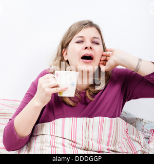 Junge Frau mit Tasse Kaffee im Bett Gähnen. Stockfoto